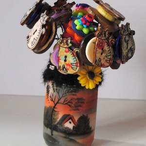 May include: A glass vase with a painted scene of a house and trees at sunset. The vase is filled with a bouquet of colorful buttons, including cat-shaped buttons, flower-shaped buttons, and buttons with geometric patterns.