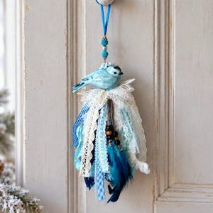 May include: A hanging ornament featuring a light blue ceramic bird perched atop a layered tassel made of white lace, blue gingham ribbon, light blue fabric strips, and dark blue feathers, suspended by a blue ribbon with small blue beads.
