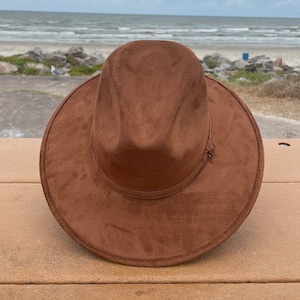 Puede incluir: Un sombrero fedora de ante marrón con ala ancha descansa sobre una superficie de madera. El sombrero tiene una banda simple alrededor de la corona y un pequeño detalle decorativo. El fondo muestra una playa y el océano bajo un cielo nublado.