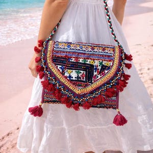 Op de afbeelding: Een kleurrijke, geborduurde schoudertas met een klepsluiting. De tas heeft ingewikkelde patronen in rood, blauw, geel en wit, geaccentueerd met rode kwastjes en een gevlochten band. De tas wordt gedragen tegen een witte jurk op een roze zandstrand.