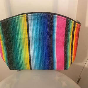Könnte beinhalten: Eine bunte, gestreifte Stofftasche mit einem schwarzen Reißverschluss. Die Tasche ist aus einem gewebten Stoff mit verschiedenen Farben, darunter Pink, Blau, Grün, Gelb und Schwarz.
