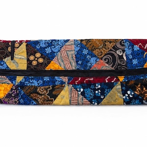 May include: A cylindrical zipped pouch featuring a patchwork design of various triangular fabric pieces in shades of blue, yellow, brown, and red. The exterior displays diverse batik-style patterns and textures with a black zip closure.