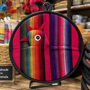May include: A round fabric pot holder featuring a multi-coloured striped pattern in shades of pink, purple, red, orange, yellow, green, and blue. A three-dimensional chicken head with a red comb and beak is attached to the front. It has a black trim.