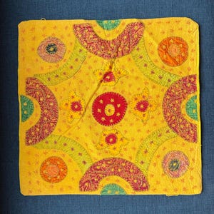 May include: Square yellow textile with embroidered floral and geometric patterns in red, green, and orange. The design includes circles, arcs, and star-like accents, creating a vibrant, handcrafted appearance. The textile is likely a decorative pillow cover or wall hanging.