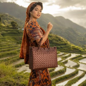Può includere: Una borsa tote in pelle intrecciata marrone con due manici. La borsa è tenuta da una persona che indossa un abito arancione fantasia e un copricapo. Lo sfondo presenta risaie terrazzate e montagne.