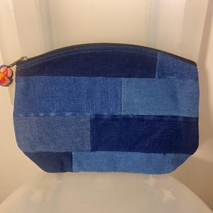 May include: A blue denim patchwork makeup bag with a zipper closure. The bag is made of different shades of blue denim and has a small, colourful tassel.