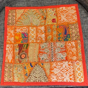 May include: A square, patchwork textile with an orange and red color scheme. The design features various patterns, including paisley and floral motifs, with gold and silver embellishments. The edges are trimmed with a bright red border.