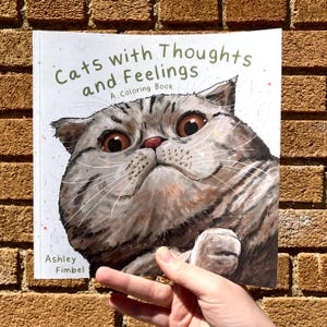 Puede incluir: Un libro para colorear titulado "Gatos con pensamientos y sentimientos: Un libro para colorear" que presenta un primer plano de la cara de un gato con ojos grandes y expresivos.