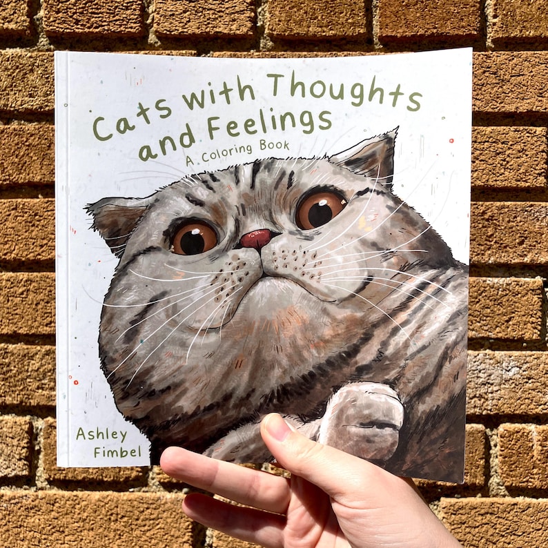 Puede incluir: Un libro para colorear titulado "Gatos con pensamientos y sentimientos: Un libro para colorear" que presenta un primer plano de la cara de un gato con ojos grandes y expresivos.