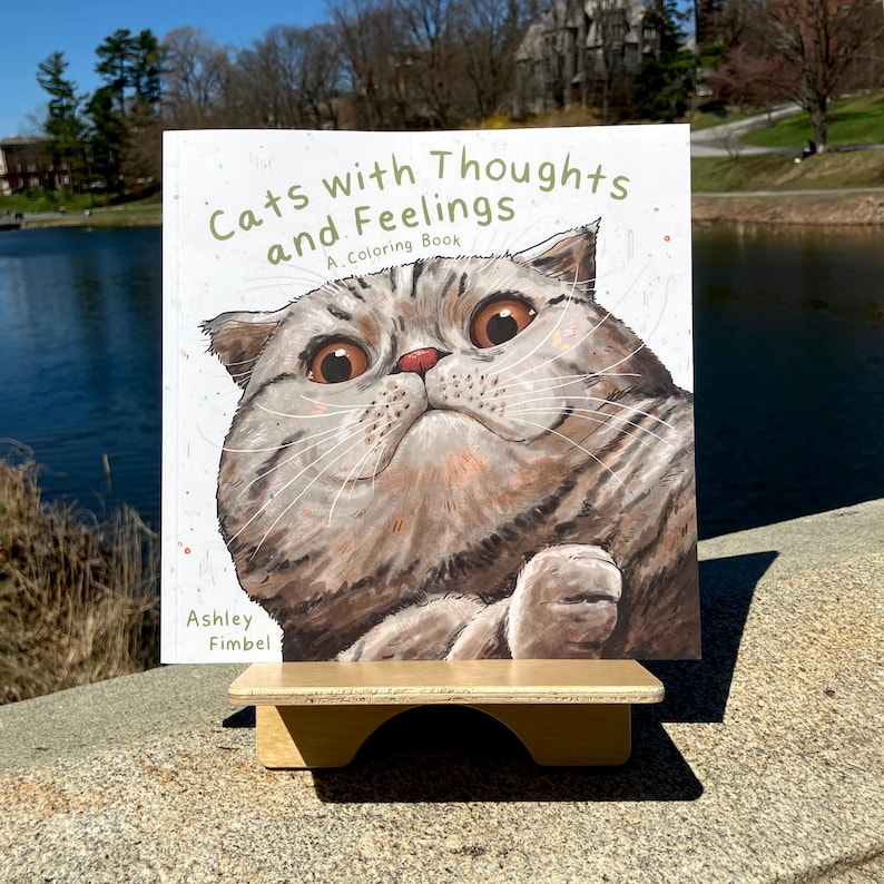 Puede incluir: Un libro para colorear titulado "Gatos con pensamientos y sentimientos: Un libro para colorear" que presenta un primer plano de un gato con ojos grandes y expresivos. El gato est&aacute; mirando directamente al espectador. El libro es de Ashley Fimbel.