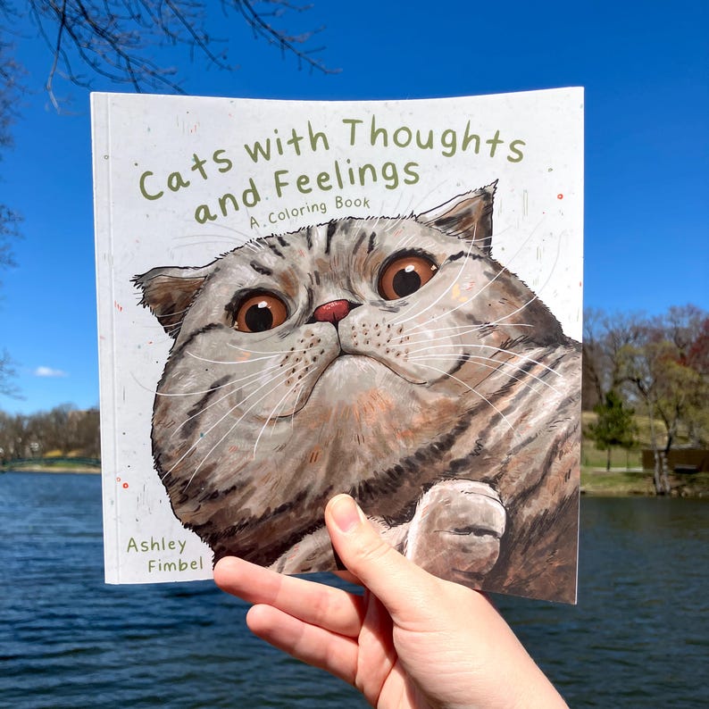 Puede incluir: Un libro para colorear titulado "Gatos con pensamientos y sentimientos: Un libro para colorear" que presenta un primer plano de la cara de un gato con ojos grandes y expresivos.