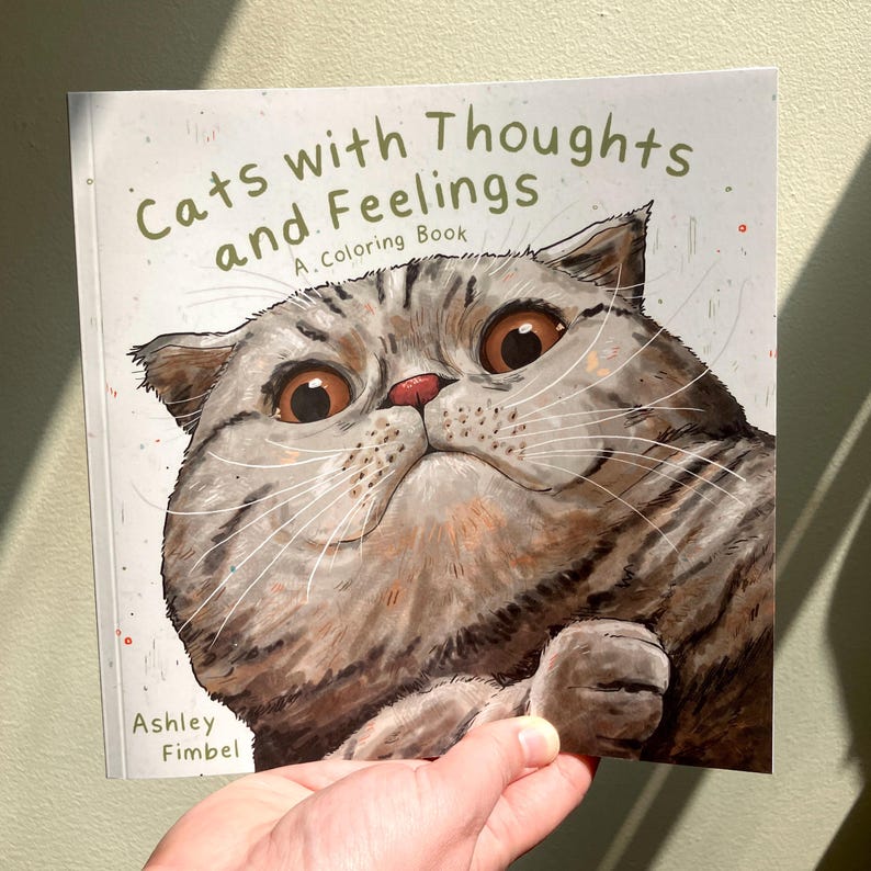 Puede incluir: Un libro para colorear titulado "Gatos con pensamientos y sentimientos: Un libro para colorear" de Ashley Fimbel. La portada presenta una ilustraci&oacute;n de primer plano de un gato gris y marr&oacute;n con grandes ojos expresivos.