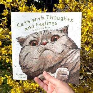 Puede incluir: Un libro para colorear titulado "Gatos con pensamientos y sentimientos: Un libro para colorear" de Ashley Fimbel. La portada presenta una ilustraci&oacute;n de primer plano de un gato gris y blanco con grandes ojos expresivos.