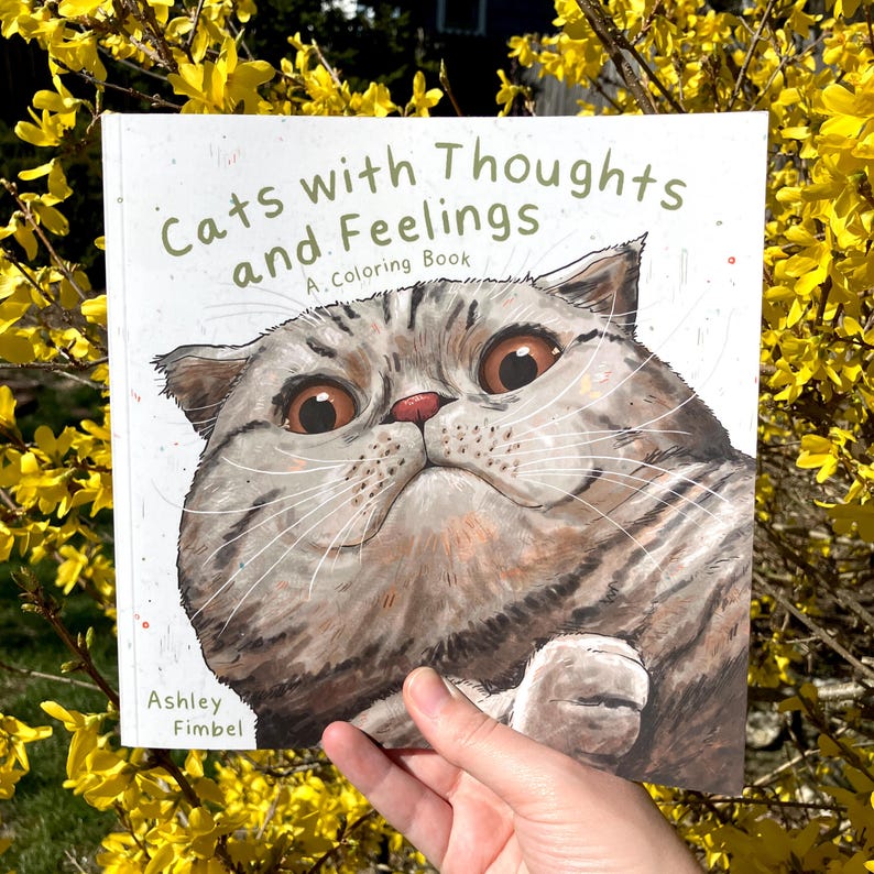 Puede incluir: Un libro para colorear titulado "Gatos con pensamientos y sentimientos: Un libro para colorear" de Ashley Fimbel. La portada presenta una ilustraci&oacute;n de primer plano de un gato gris y blanco con grandes ojos expresivos.