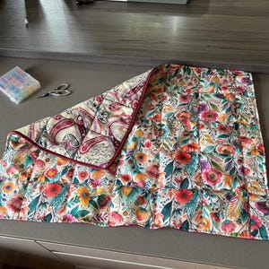 Könnte beinhalten: Ein gestepptes Stoffstück mit einem lebendigen Blumenmuster aus roten, orangen, gelben und grünen botanischen Elementen auf der einen Seite und einer cremefarbenen Rückseite mit dunkelrotem und schwarzem Paisle und schwarzem Paisleymuster, eingefasst mit dunkelroter Borte.