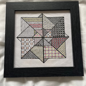 Könnte beinhalten: Gerahmtes Kreuzstich-Kunstwerk mit einem geometrischen Sternmuster. Das Design beinhaltet verschiedene Muster und Farben, darunter Schwarz, Weiß, Rot und Gelb. Das Kunstwerk wird in einem schwarzen Holzrahmen präsentiert.