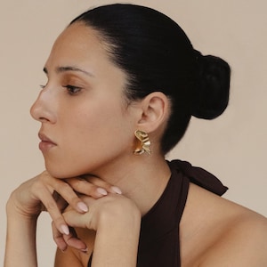 Puede incluir: Un pendiente de tono dorado con un diseño curvo, ondulado o con volantes. El pendiente tiene un acabado metálico pulido y una forma estructurada y plegada que refleja la luz, colocado en el lóbulo de la oreja con un cierre seguro.