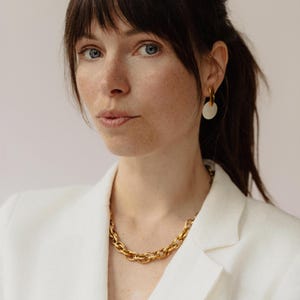 Könnte beinhalten: Eine Frau trägt eine goldene Kette und goldene Creolen mit einem weißen, kreisförmigen Anhänger. Sie trägt einen weißen Blazer. Der Schmuck ist der Blickfang des Bildes.