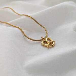 Könnte beinhalten: Eine goldfarbene Halskette mit einem Brezel-Anhänger. Der Anhänger hat eine stilisierte Brezelform, und die Kette ist in einem dünnen, schlangenartigen Design gehalten. Die Halskette wird auf einem weißen, strukturierten Stoff präsentiert.