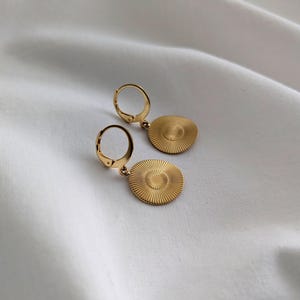 Può includere: Un paio di orecchini color oro. Ogni orecchino presenta un pendente circolare con un design a raggi di sole. Gli orecchini hanno una chiusura a leva e sono esposti su una superficie bianca strutturata.