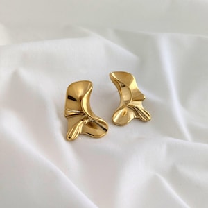 Op de afbeelding: Een paar goudkleurige oorstekers. Elke oorbel heeft een uniek, abstract ontwerp met een gebogen, gevouwen uiterlijk. De oorbellen worden gepresenteerd op een witte, getextureerde stof, die hun metallic glans benadrukt.