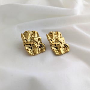 May include: A pair of gold-coloured stud earrings with a textured finish. The earrings have an abstract, crumpled design, reflecting light. They are presented against a white backdrop.