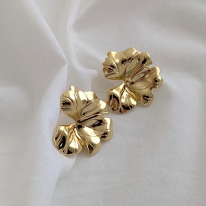 Puede incluir: Un par de pendientes de hoja de oro. Los pendientes son grandes y tienen una superficie texturizada.