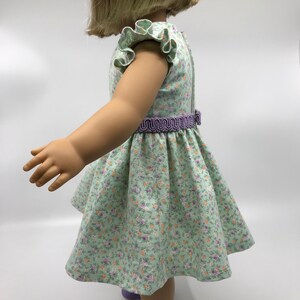 May include: A green floral dress with a purple ruffle trim on the sleeves and a purple elastic waistband. The dress is on a doll with blonde hair.