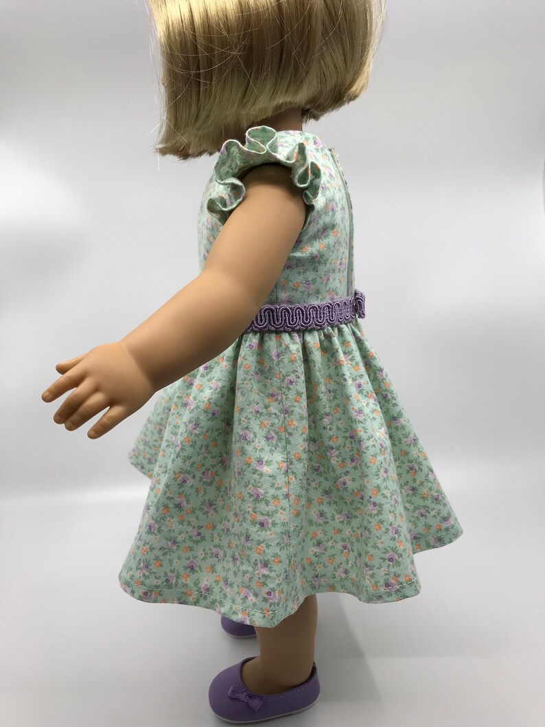 May include: A green floral dress with a purple ruffle trim on the sleeves and a purple elastic waistband. The dress is on a doll with blonde hair.