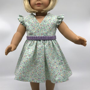 May include: A green floral print dress with a purple ribbon belt and short sleeves. The dress is worn by a doll with blonde hair and blue eyes.