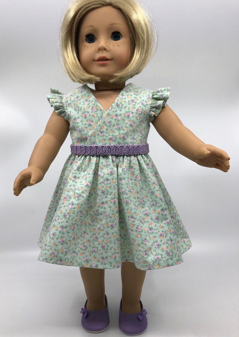 May include: A green floral print dress with a purple ribbon belt and short sleeves. The dress is worn by a doll with blonde hair and blue eyes.