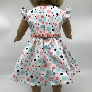 May include: A white dress with a pink belt and colorful polka dots. The dress has short sleeves with ruffles and a full skirt. The doll is wearing white shoes.