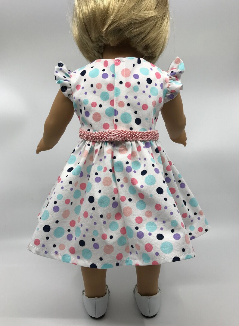 May include: A white dress with a pink belt and colorful polka dots. The dress has short sleeves with ruffles and a full skirt. The doll is wearing white shoes.