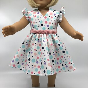 May include: A white dress with a pink, blue, and purple polka dot pattern. The dress has a V-neckline and short sleeves with ruffles. A pink braided belt is worn around the waist.