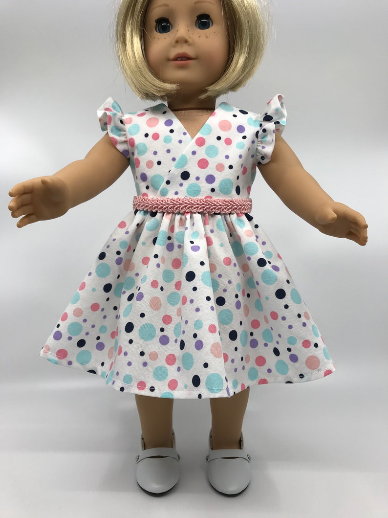 May include: A white dress with a pink, blue, and purple polka dot pattern. The dress has a V-neckline and short sleeves with ruffles. A pink braided belt is worn around the waist.