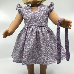May include: A purple floral dress with a V-neck and short sleeves. The dress has a gathered skirt and is worn by a doll. The doll is also wearing purple shoes and a purple headband.