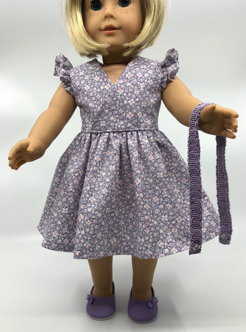 May include: A purple floral dress with a V-neck and short sleeves. The dress has a gathered skirt and is worn by a doll. The doll is also wearing purple shoes and a purple headband.