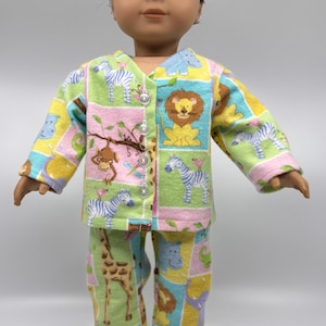 May include: A doll wearing a colorful pajama set with a jungle animal print. The set includes a button-down top and pants, featuring zebras, lions, giraffes, and monkeys in a patchwork design. The pajamas are made of a soft fabric.