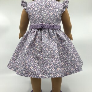 May include: A purple and white floral dress with a purple ribbon waistline. The dress is made for a doll and has short sleeves with ruffles.