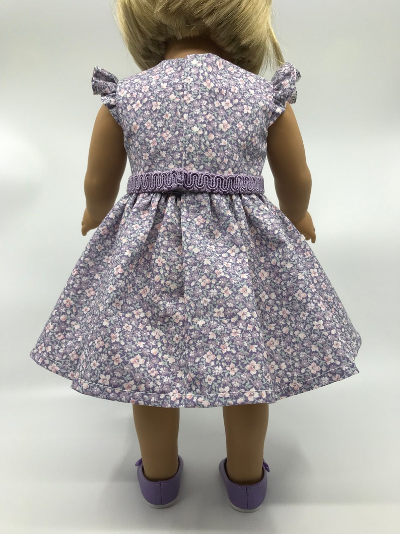 May include: A purple and white floral dress with a purple ribbon waistline. The dress is made for a doll and has short sleeves with ruffles.
