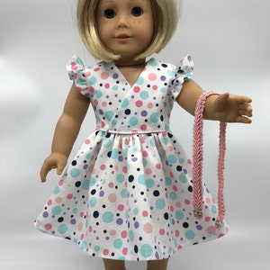 May include: A doll wearing a white dress with pink, blue, and purple polka dots. The dress has a V-neckline and ruffled sleeves. The doll is also wearing white shoes and a pink necklace.