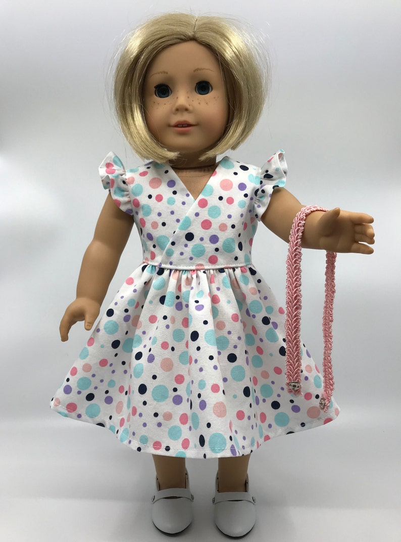 May include: A doll wearing a white dress with pink, blue, and purple polka dots. The dress has a V-neckline and ruffled sleeves. The doll is also wearing white shoes and a pink necklace.
