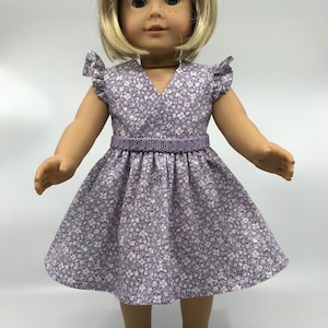 May include: A purple and white floral dress with a ruffled neckline and a purple ribbon belt. The dress is worn by a doll with blonde hair and blue eyes.
