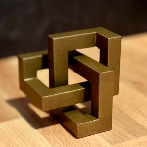 May include: A gold-colored metal sculpture of an interlocking knot design. The sculpture is made of multiple squares that are connected to each other in a complex pattern.