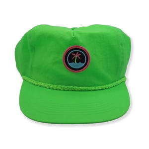 Op de afbeelding: Een felgroene baseballpet met een geborduurd palmboom op de voorkant. De palmboom is omgeven door een cirkel met een roze rand en een blauw waterontwerp.