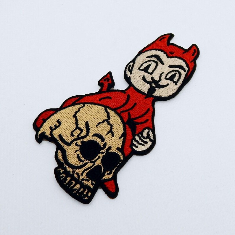 May include: Embroidered patch depicting a red devil character with a playful expression, sitting on a tan skull. The devil has horns and a pointed tail. The patch is designed with black and tan details.
