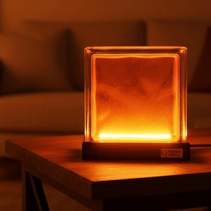 May include: A rectangular lamp with a clear glass block design, emitting warm orange light. The lamp sits on a dark wooden table, with a black base and a logo on the front. The background includes a sofa, artwork, and a potted plant.