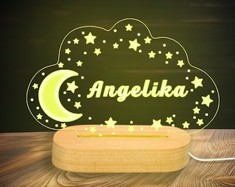May include: A night light shaped like a cloud, with the name "Angelika" illuminated in yellow. The cloud design includes stars and a crescent moon. It sits on a wooden base and is connected to a white cord.