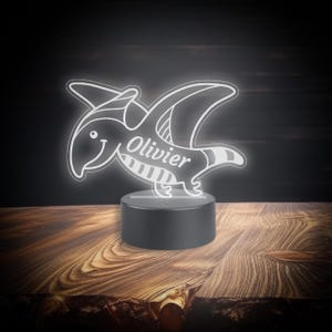 May include: A white 3D printed night light shaped like a cartoon pterodactyl with the name "Olivier" printed on its body. The night light is on a black base and is illuminated.