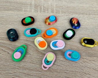 Könnte beinhalten: Sortiment an bunten Handygriffen mit verschiedenen Designs und Farben. Die Griffe sind aus Kunststoff und haben eine runde Form. Sie sind so konzipiert, dass sie an der Rückseite eines Handys befestigt werden, um einen besseren Halt zu gewährleisten.